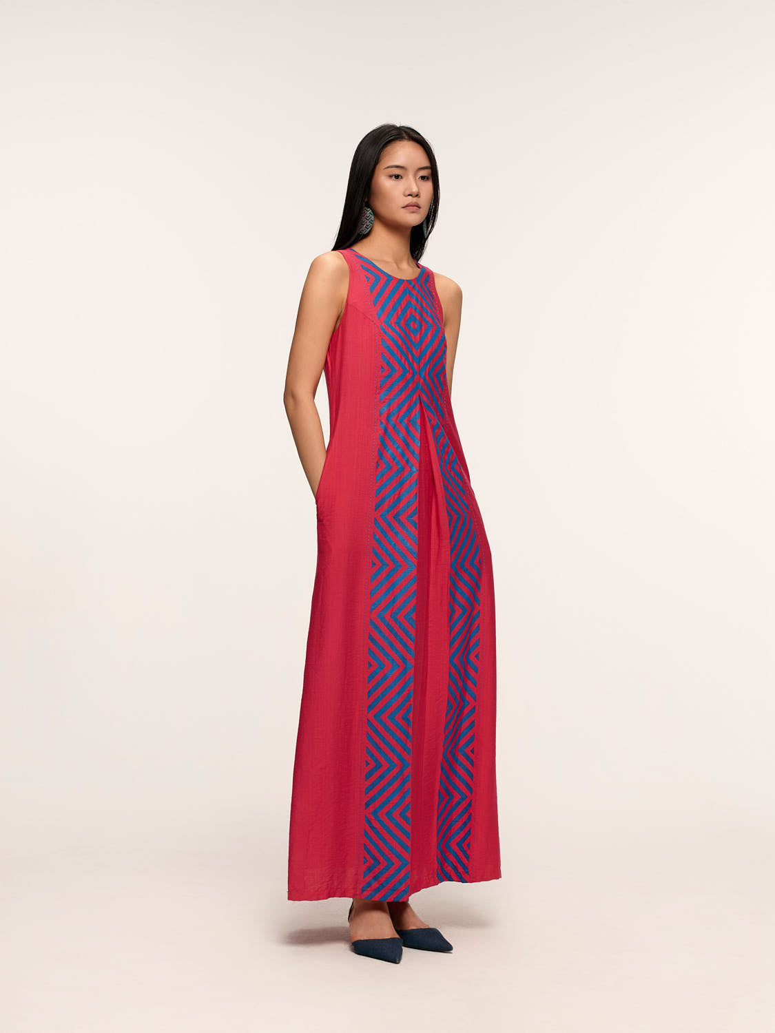 A model wearing a long pink dress with geometric patterns and embroidery details, standing against a white background.
