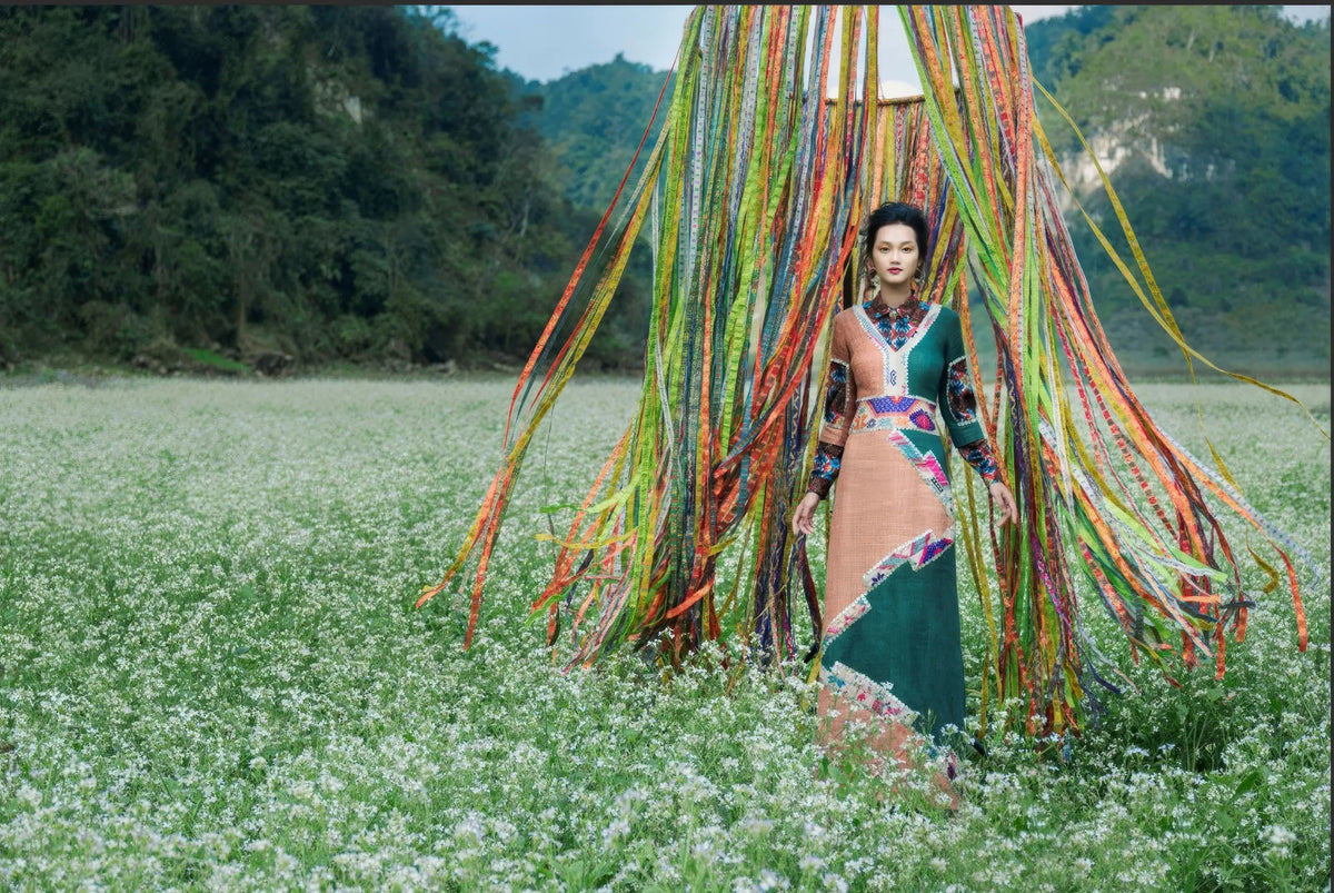 A model in a Chula patchwork áo dài stands beneath cascading ribbons in a field of white buckwheat flowers in northern Vietnam