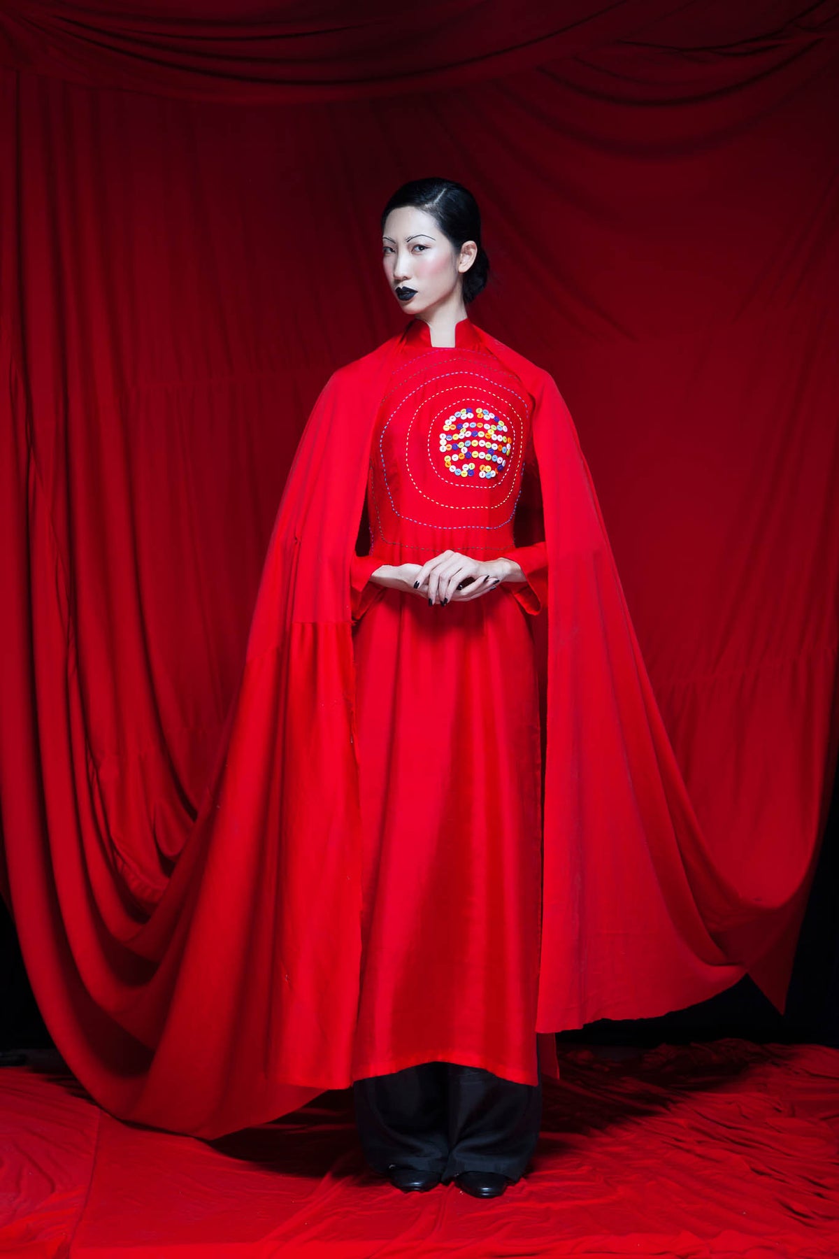 Model in a red cape and ao dai painted with a longevity character, against a red curtain