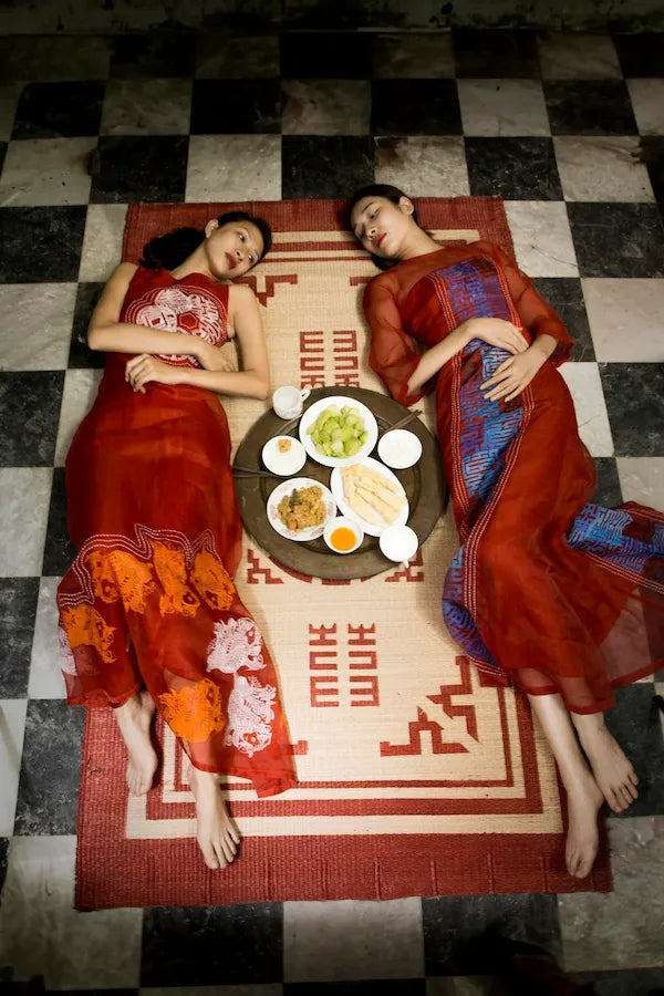 Two models in red silk ao dai lying head to head on a woven mat around a tray of food, on a chequered floor
