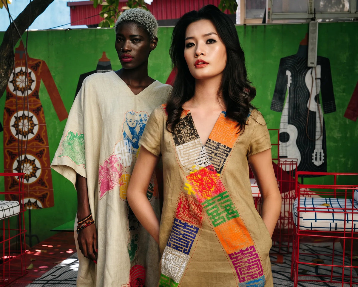Two models in natural linen pieces patched with hand-painted panels, before a green wall hung with Chula artwork