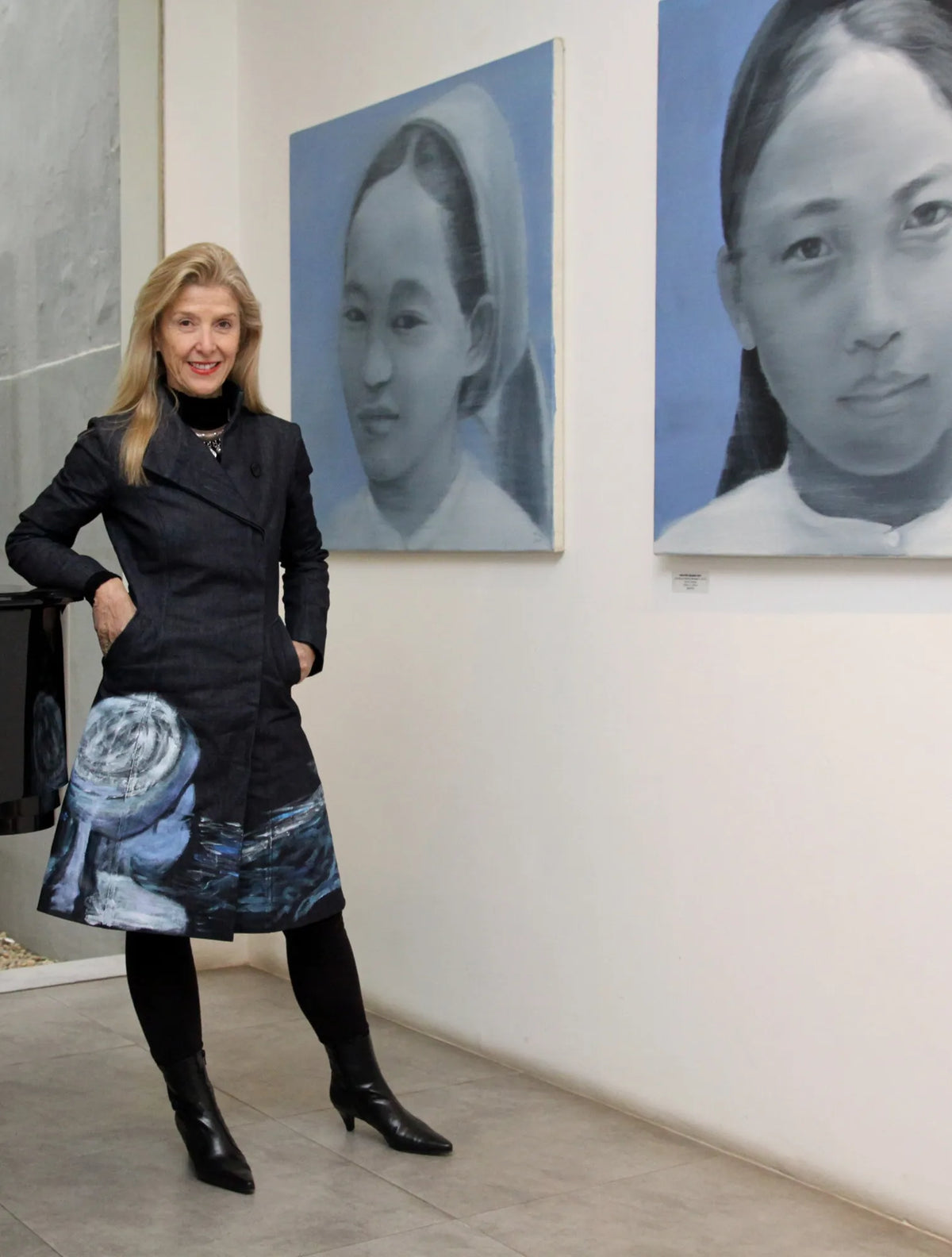 Suzanne Letch in a Chula coat between two portrait paintings at Art Vietnam Gallery